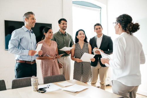 A group of six diverse business professionals are standing in a bright, modern office. They are smiling and holding papers while engaging in a discussion. One person, facing away from the camera, appears to be leading the meeting. A table with documents and a coffee cup is in the foreground, and a large screen is mounted on the wall behind the group. Natural light fills the room through a window.