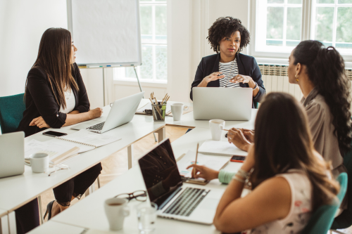 Four women in a professional meeting setting, seated around a table with laptops, notebooks, and coffee cups.