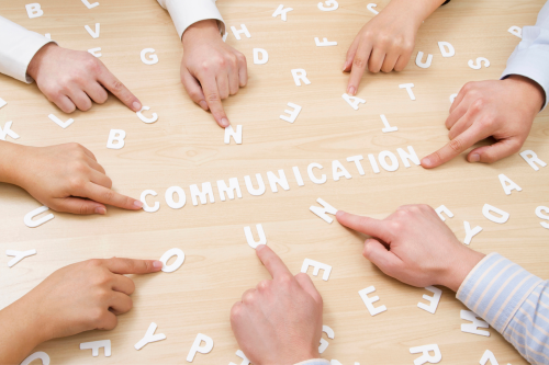 A group of hands pointing towards the word "COMMUNICATION" spelled out with individual letters on a wooden table.