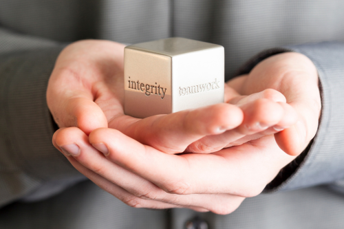 The image shows a close-up of a person wearing a suit, holding a metallic cube with the words "integrity" and "teamwork" engraved on its sides.