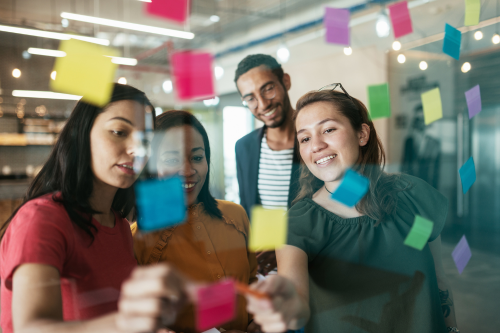 A group of four diverse professionals collaborating in an office, standing around a glass board covered with colorful sticky notes